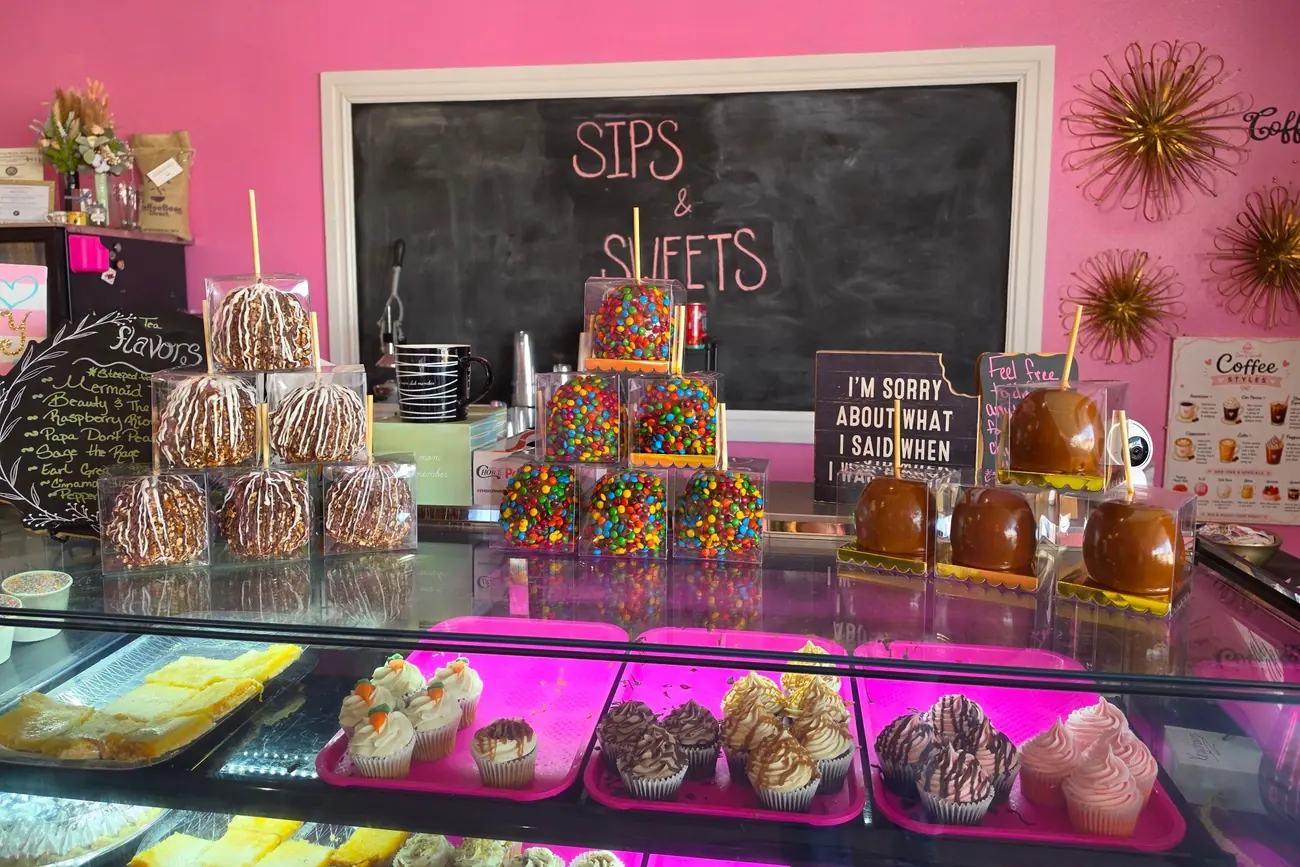 Inside the shop: candy-covered apples stacked on the counter above a lit case of cupcakes, with the chalkboard and pink wall behind