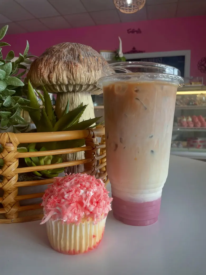 An iced latte and a pink coconut cupcake on a table inside Sips & Sweets, with the pink wall behind